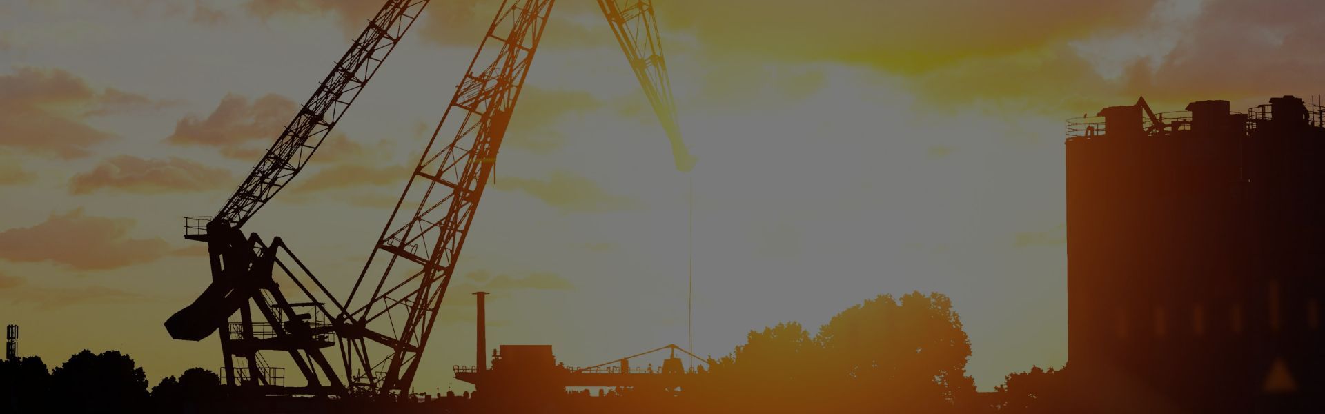 A silhouette of two lattice crane booms against a sunset sky with low industrial buildings and trees along the hori
zon